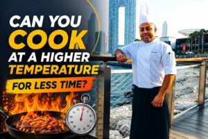 Chef in a white uniform standing by a waterfront railing with a modern city skyline behind him, next to bold text reading “Can You Cook at a Higher Temperature for Less Time?” with flames, a sizzling steak, and a stopwatch graphic emphasizing fast, high-heat cooking.