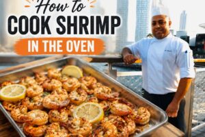 A professional chef in a white uniform stands on a waterfront boardwalk with a modern city skyline in the background. In the foreground, a tray of golden-brown roasted shrimp garnished with lemon slices is prominently displayed. Large bold text reads, “How to Cook Shrimp in the Oven,” making the image a featured graphic for a shrimp cooking tutorial.