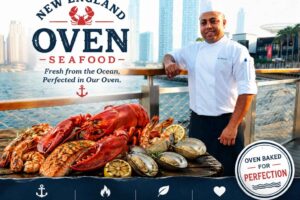 Promotional seafood restaurant feature image showing a chef in a white uniform standing by a waterfront with a city skyline in the background. The design features “New England Oven Seafood” branding, a fresh seafood platter with lobster, shrimp, salmon, clams, and lemon slices, plus text highlighting fresh ocean seafood, oven perfection, quality ingredients, and passion. A nautical-inspired restaurant advertisement with a premium coastal dining theme.